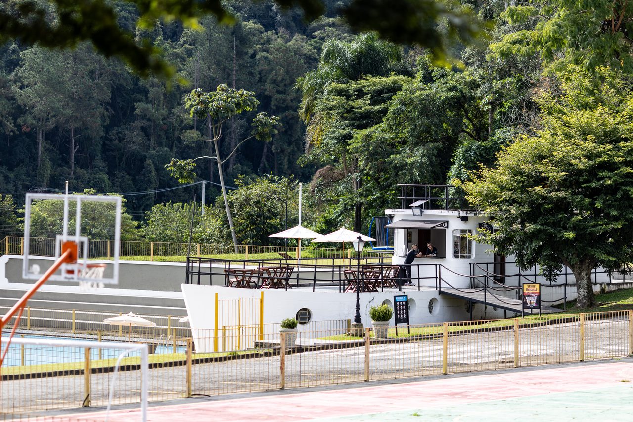 Bar em formato de barco ao lado da piscina, com a quadra de basquete e a mata ao fundo