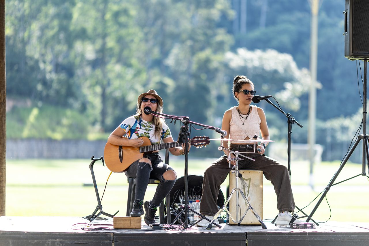 Dupla tocando violão e percussão no palco ao ar livre, em tarde de música ao vivo