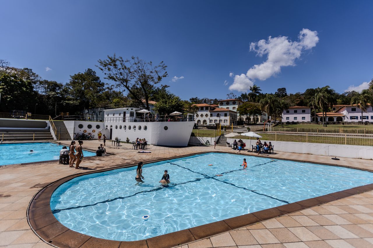 Piscina do Instituto da Costela com pessoas na água em dia de céu azul, com os casarões e a serra ao fundo