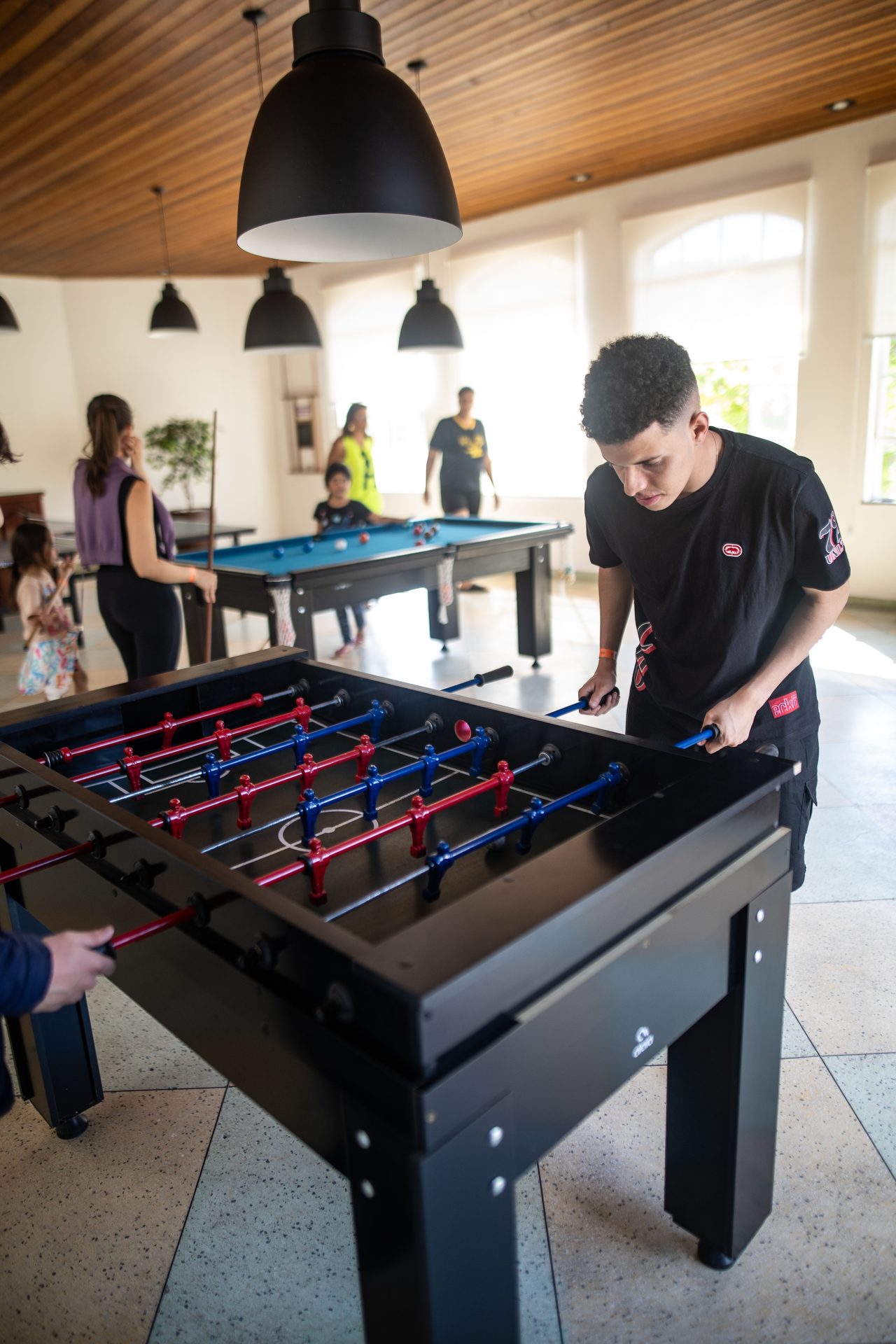 Pessoas jogando pebolim no salão de jogos coberto, com a mesa de sinuca ao fundo