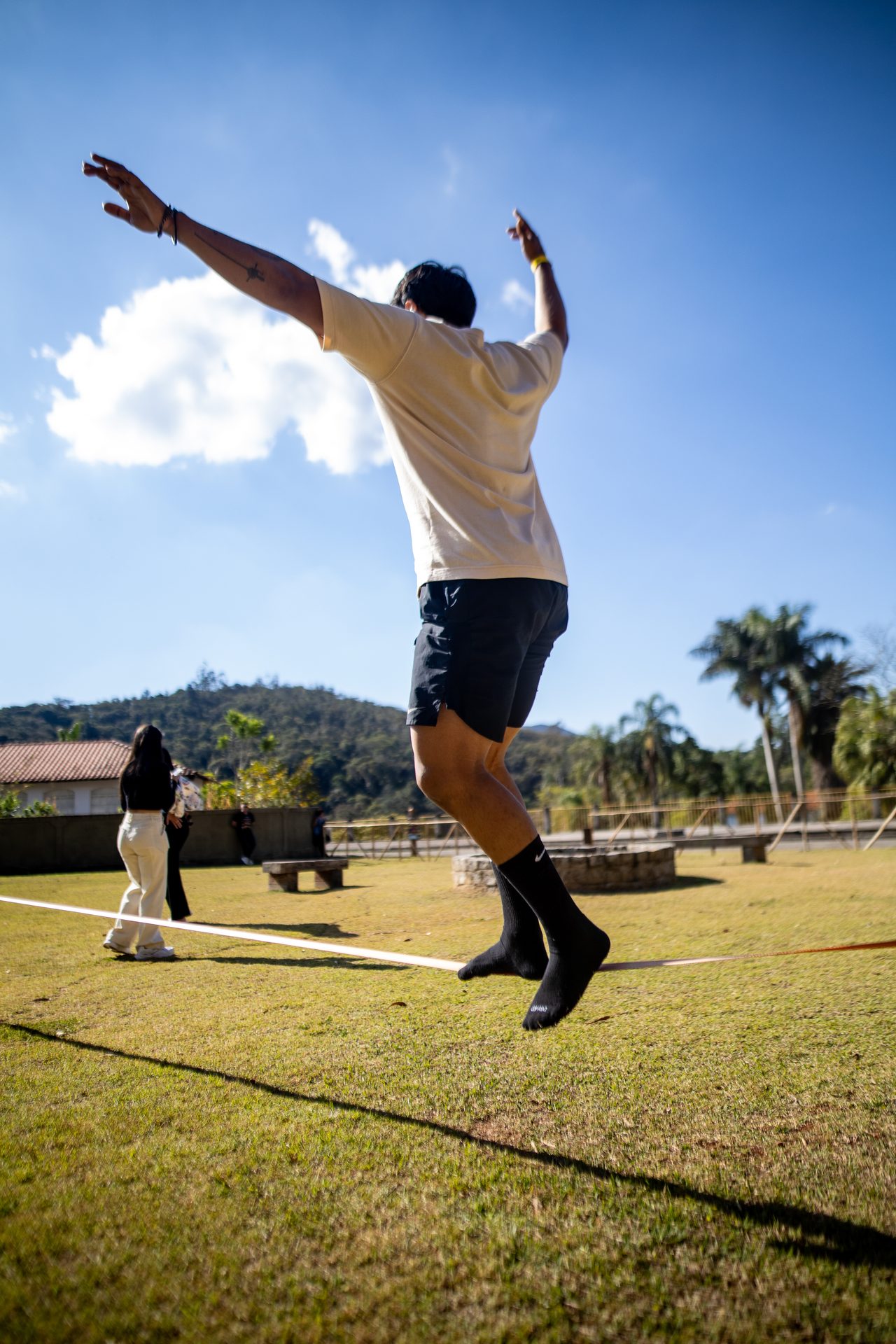 Visitante saltando na fita de slack line, com o céu azul e as palmeiras ao fundo